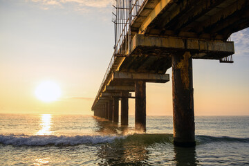 Ocean waves beneath industrial pier under golden sun, decay and beauty captured single marine frame. Pier skeleton extends into calm sea, scenic sunset light casts shadows on rippling water, Bulgaria