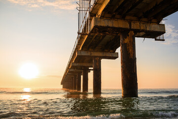 Abandoned pier stretching into calm Black Sea at golden sunrise, rusty structure reflecting on gentle waves in Bulgaria. Rusted metal construction old pier leads into ocean horizon during golden hour