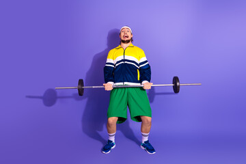 Young man lifting a barbell with funny expression against a solid purple background wearing retro...