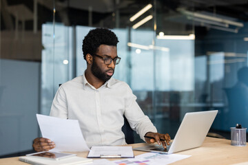 An African American businessman wearing glasses works on a laptop in a modern office setting,...