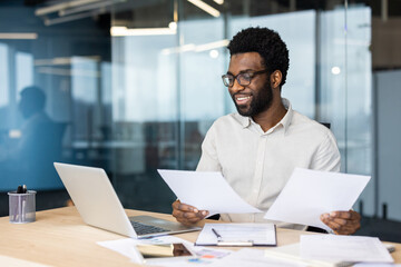 An African American man smiles while reviewing documents at his desk in a modern office setting,...