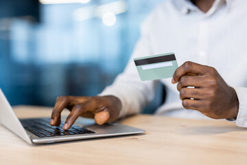 An individual, with dark skin, is using a laptop and holding a credit card, possibly for online...