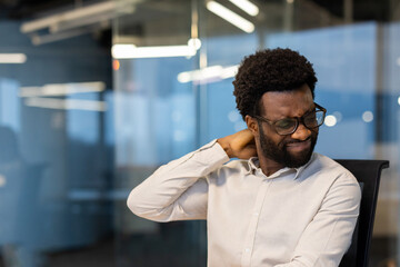 A man with glasses touches his neck, appearing to suffer from neck pain or strain while sitting in...