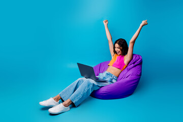 Joyful young woman on beanbag celebrating success with laptop against vibrant blue background in...