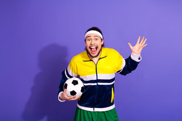 Young energetic athlete wearing colorful retro sportswear holding a soccer ball joyfully on a purple background