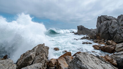 Fototapeta premium ocean waves crashing against jagged rocks at the edge of a coast, white foam and mist flying into the air, cloudy sky above.
