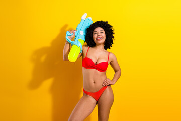 Cheerful woman in vibrant red bikini posing with a water gun against a bright yellow background, exuding joy and summer vibes.