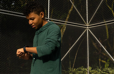 Dark skinned Latino teenager looks at his smartwatch while exercising in a park in the morning, outdoor lifestyle