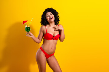 Joyful young woman in red bikini holding a cocktail, vibrant yellow background, confident and...