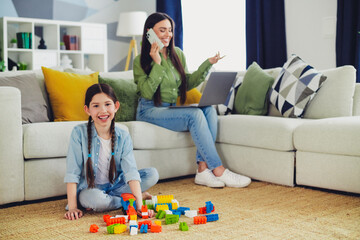 Mother multitasking with laptop while her daughter plays at home, enjoying quality family time with...