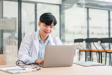 Young male doctor wearing white coat working on laptop in modern office, smiling and focused, with stethoscope and clipboard on desk, natural light from large windows