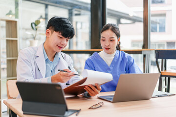 Two young Asian medical professionals, man and woman, wearing lab coats and scrubs, collaborate at desk with laptop and clipboard in modern office, showing teamwork and focus