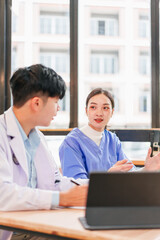 Two young Asian medical professionals, man and woman, wearing lab coats and scrubs, discuss patient care at table with laptop in bright hospital office