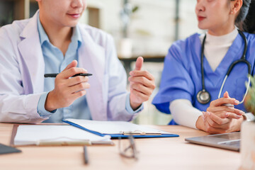 Two healthcare professionals, one in white coat and one in blue scrubs, discuss patient information at desk with documents and laptop, showing teamwork and focus in medical office
