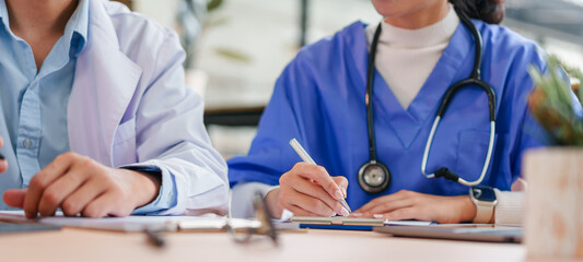 Two healthcare professionals, one in white coat and one in blue scrubs, discuss patient information at desk with documents and laptop, showing teamwork and focus in medical office