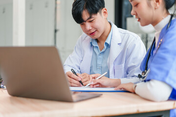 Young Asian male and female medical professionals in lab coats working together at desk, discussing documents and using laptop, showing teamwork and focus in clinical setting
