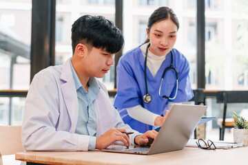 Two young Asian medical professionals, male doctor and female nurse, collaborate at desk with laptop in bright hospital office, discussing patient care with focused expressions