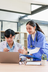 Two healthcare professionals, one in white coat and one in blue scrubs, discuss patient information at desk with documents and laptop, showing teamwork and focus in medical office