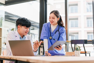 Young Asian male doctor and female nurse in blue scrubs discussing patient information with digital tablet and laptop, smiling and collaborating in modern hospital office