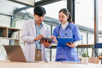 Two Asian medical professionals, male doctor white coat and female nurse blue scrubs with stethoscope, discuss patient information using digital tablet and clipboard bright hospital office