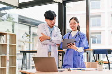 Two healthcare professionals, one male and one female, stand in modern medical office discussing patient information on digital tablet, appearing focused and collaborative