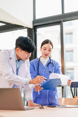 Two Asian medical professionals, male doctor and female nurse, discuss patient information with focus and teamwork in bright hospital office environment