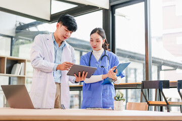 Two Asian medical professionals, male doctor and female nurse, discuss patient information with focus and teamwork in bright hospital office environment