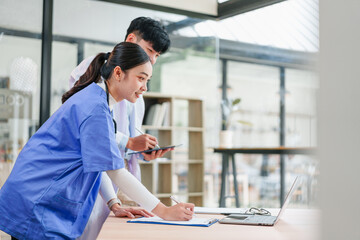 Two young Asian medical professionals in blue and white uniforms collaborate in bright modern hospital office, using laptop and clipboard, focused and engaged in teamwork