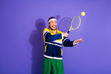 Young energetic male tennis player in stylish retro sportswear playing in front of a purple studio...