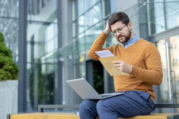 Shocked young male businessman sitting on a bench outside, holding a laptop on his lap, reading a...