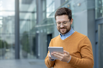 Close-up photo of a young smiling man standing outside and using a tablet in his hands