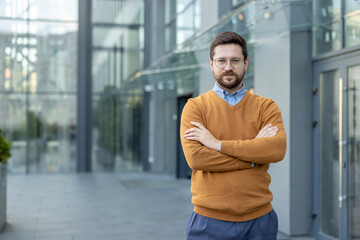 Portrait of a confident businessman, developer standing outside an office building, arms crossed on his chest and looking at the camera