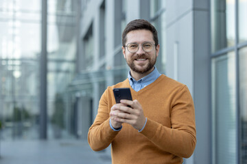 Portrait of a young smiling businessman standing on the street, holding a phone and looking at the...