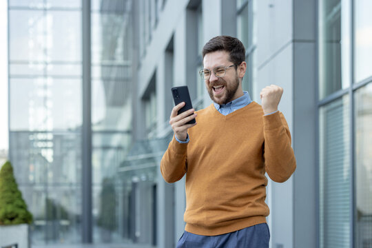 Happy young male businessman standing near the building, looking at the phone screen and rejoicing in success