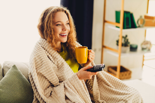 Young woman enjoying a cozy moment on a sofa, holding a cup of tea while watching TV in a comfortable home setting