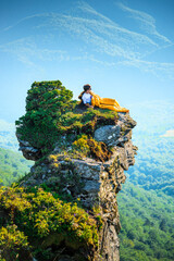 Obraz premium Man relaxing in sleeping bag on mountain cliff, enjoying scenic sunrise view over forested hills
