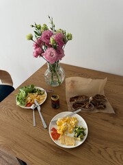Two breakfast plates with eggs, cheese, avocado, and vegetables on a wooden table. Sliced bread and a vase with flowers are also visible.