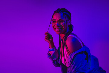 Charming young woman with trendy glasses and braided hairstyle, enjoying vibrant neon lights in a...