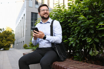 Successful businessman with smartphone on bench near office building