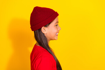 Side profile of cheerful young girl in red sweater and beanie against a bright yellow background, smiling vividly