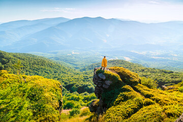 Man wrapped in a blanket standing on mountain cliff, admiring panoramic view of forest valley and distant hills at sunset