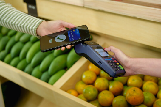 Closeup hands holding mobile phone above POS machine completing digital payment inside fruit shop concept of contactless sale in small business modern grocery store with fresh tropical produce - Powered by Adobe