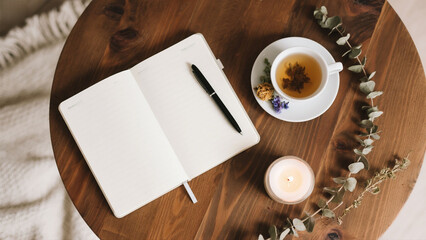 A tranquil scene of a blank notebook, pen, teacup, candle, and eucalyptus on a wooden table.
