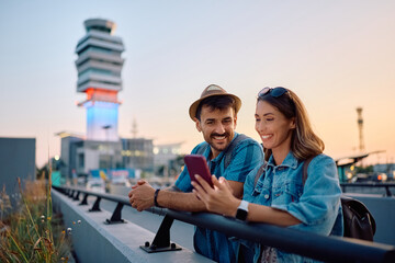 Happy couple of tourists using cell phone at the airport at sunset.