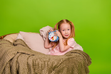Cheerful young girl holding an alarm clock in a cozy blanket against a vibrant green background