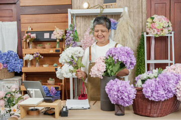 Senior asian florist behind shop counter arranging multiple artificial flowers into black vase with...