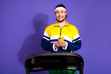 Cheerful young man in colorful sportswear on treadmill holding smartphone against a purple...