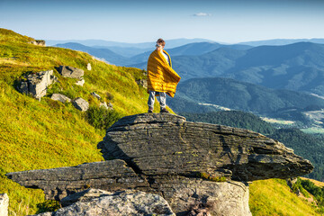 Man wrapped in a blanket standing on mountain cliff, admiring panoramic view of forest valley and distant hills at sunset