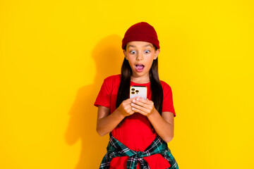 Young girl in red t-shirt and beanie expressing amazement while holding smartphone on bright yellow background