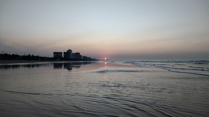 Nascer do sol dourado na Praia Grande, SP. Silhuetas urbanas refletidas na orla calma sob luz...